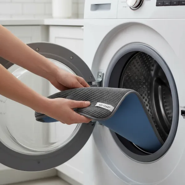 A close-up of the All-Clad reversible drying mat being placed into a washing machine, illustrating its easy care and machine washable feature.